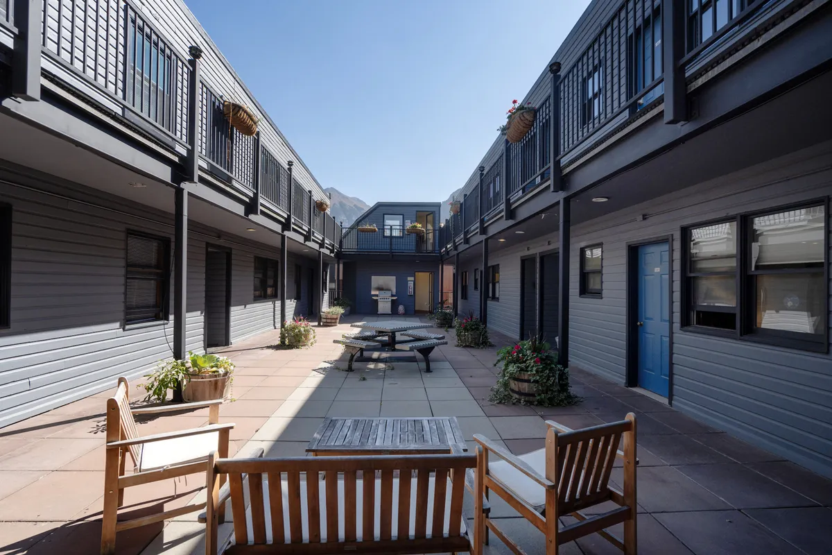 The image features a sunny courtyard with a communal seating area, planters filled with flowers, a blue door, and balconies overlooking the space. The ambiance is inviting and social, ideal for gatherings or relaxing outdoors.