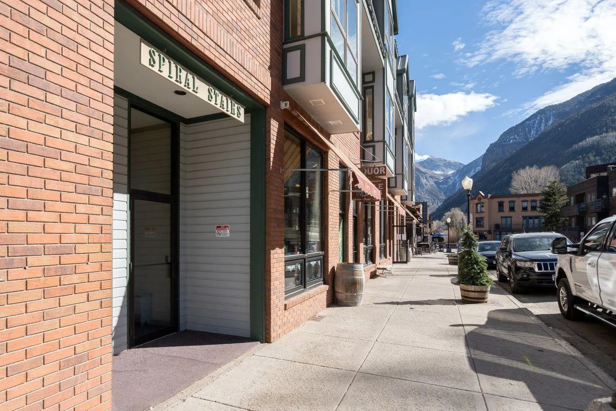 A dynamic street scene with the spiral stairs entrance, brick facade, mountain backdrop, and street furniture. Locals and visitors are seen strolling, shopping, and dining, capturing the lively, walkable atmosphere of Telluride.