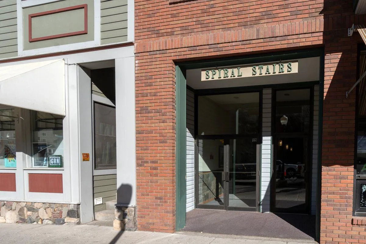The exterior entrance to the condo features a prominent 'SPIRAL STAIRS' sign, a charming brick facade, and green trim. The image conveys the convenience of being steps away from Telluride’s walkable downtown and helps establish a sense of place and accessibility.