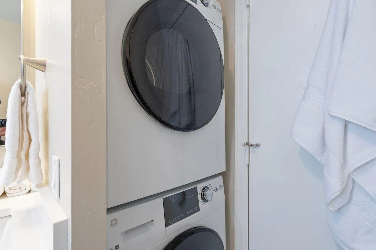 A stacked washer and dryer set is neatly placed against a wall, with towels hanging on a nearby rack. The space is bright, clean, and organized, showcasing the laundry facilities offered to residents.