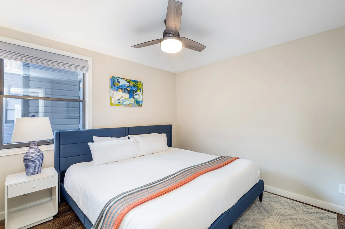 A serene bedroom with a comfortable bed, soft lighting from a bedside lamp, tasteful wall art, and a ceiling fan overhead. The space feels quiet and inviting, offering an ideal setting for relaxation and restful sleep.