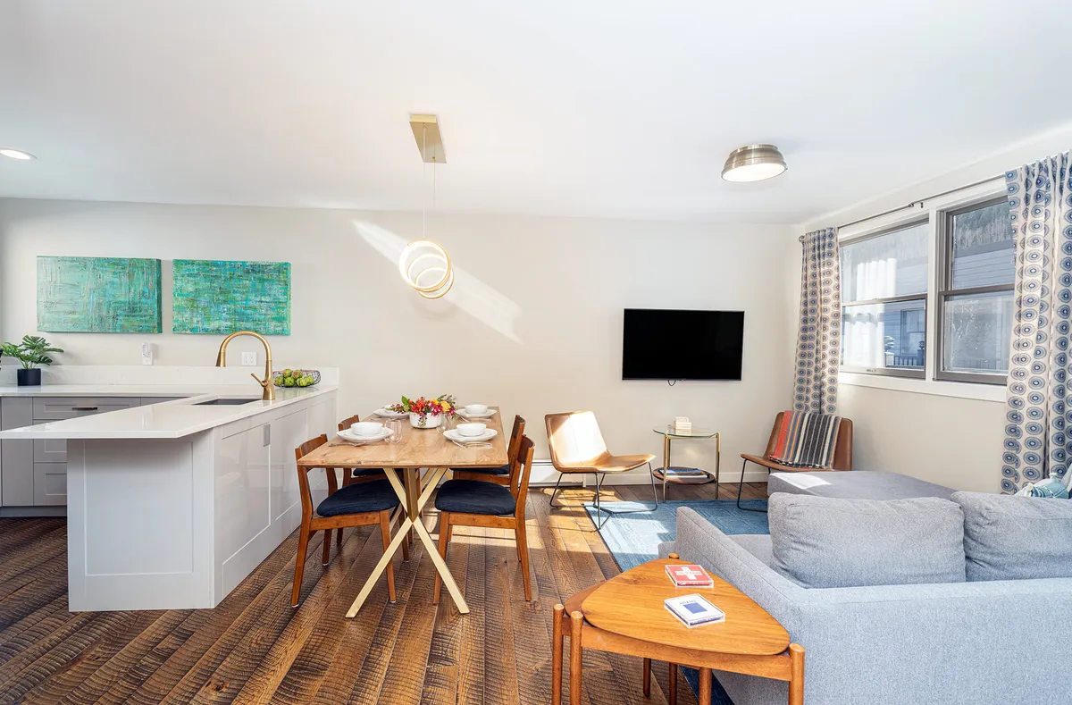 A welcoming living space featuring a dining table set for a meal, a modern kitchen island, and a comfortable seating area. Artwork and a bright, airy atmosphere add a touch of warmth and personality to the room.