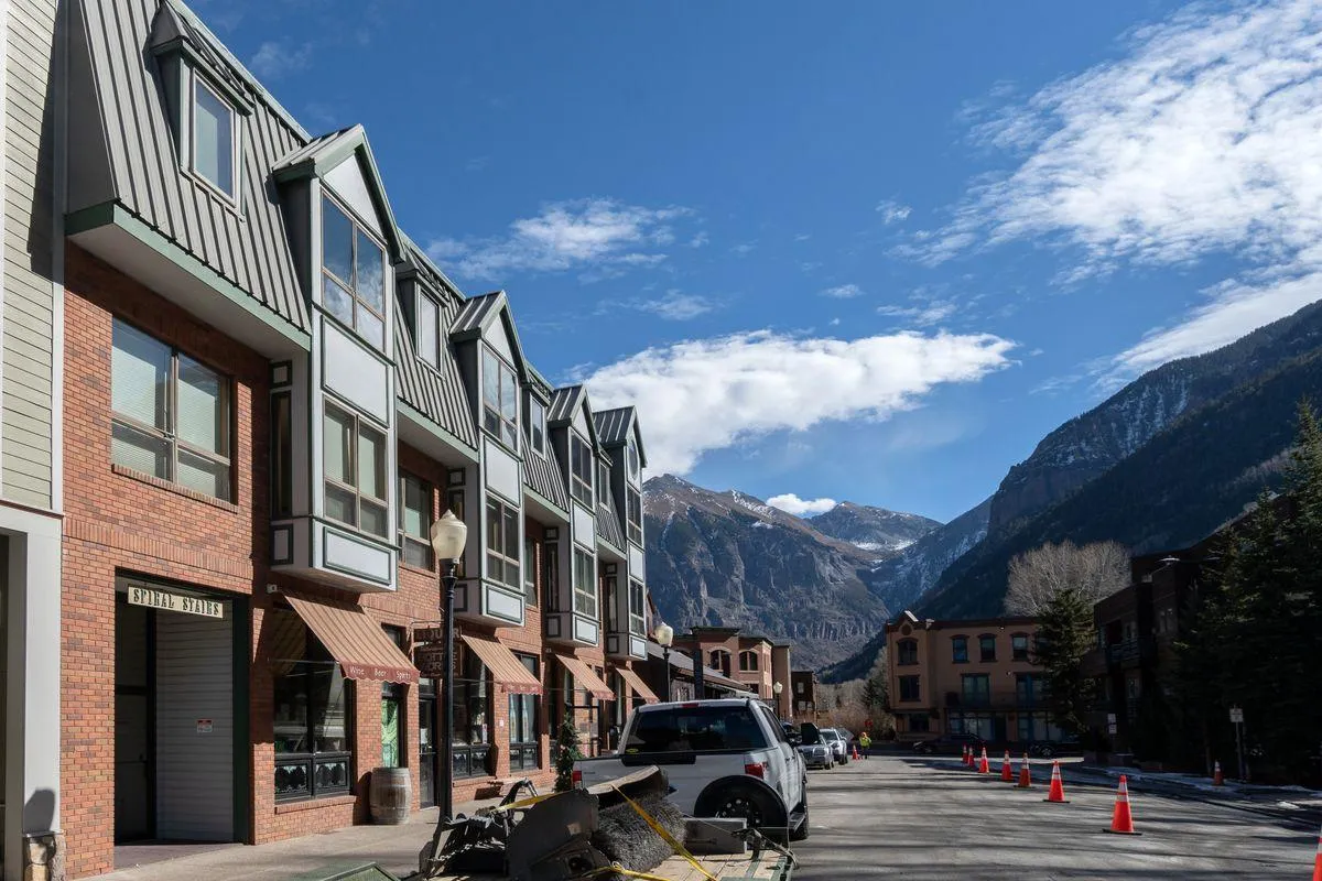 A vibrant exterior street view with charming local architecture, a mountain backdrop, and activity on the streets. People are seen walking and biking, enjoying the lively atmosphere of the town’s main drag.