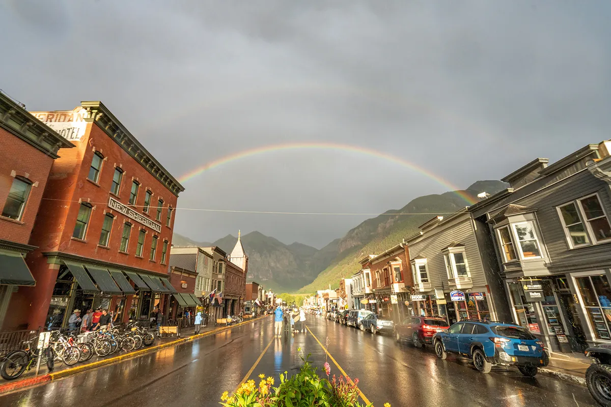 This inviting street scene features historic brick buildings adorned with bicycles and cars, a lively sidewalk, and a vibrant rainbow arching overhead. The atmosphere is bustling and full of energy, suggesting a charming, walkable downtown filled with unique shops, cafes, and local color.
