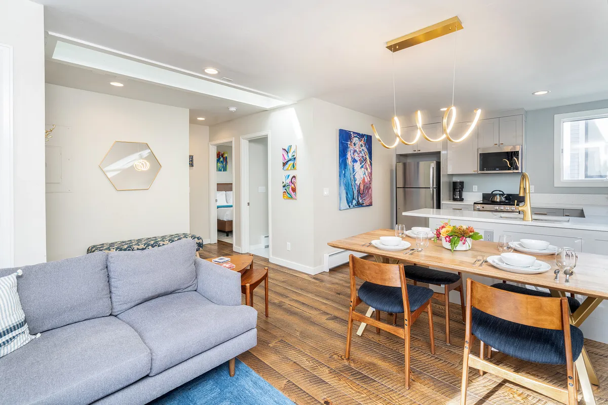 A cozy interior view showing a versatile dining table, a plush sofa, and stylish artwork on the walls. The kitchen appliances are visible in the background, tying all the main living spaces together in a single shot.
