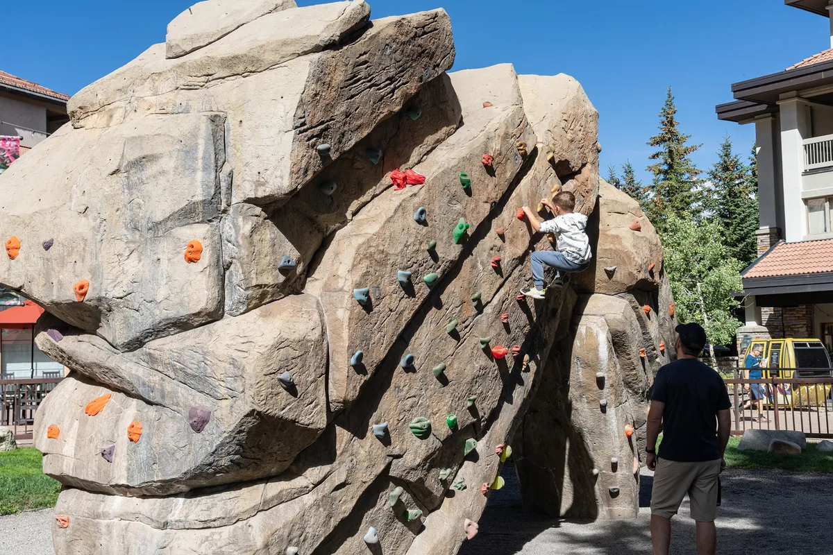 A child is climbing a rock structure under the watchful eye of an adult, set in an outdoor playground space. The scene is filled with a sense of adventure and family bonding, capturing the joy of outdoor play in a safe and inviting environment.