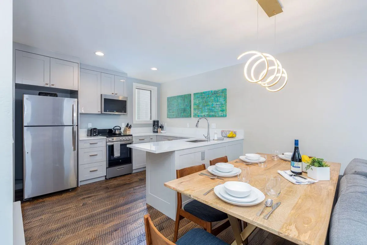 A bright, open-plan space featuring a dining table set for a meal adjacent to a modern kitchen with gleaming appliances. Abstract wall art adds a contemporary touch, while the room is suffused with warm, inviting natural light.
