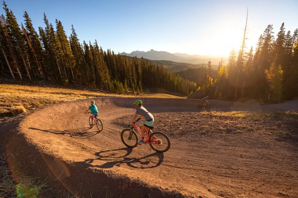 Bikers ride along a dirt path winding through a dense forest with dramatic mountains in the background. The scene captures the exhilaration of exploring Telluride’s trails during warmer months.