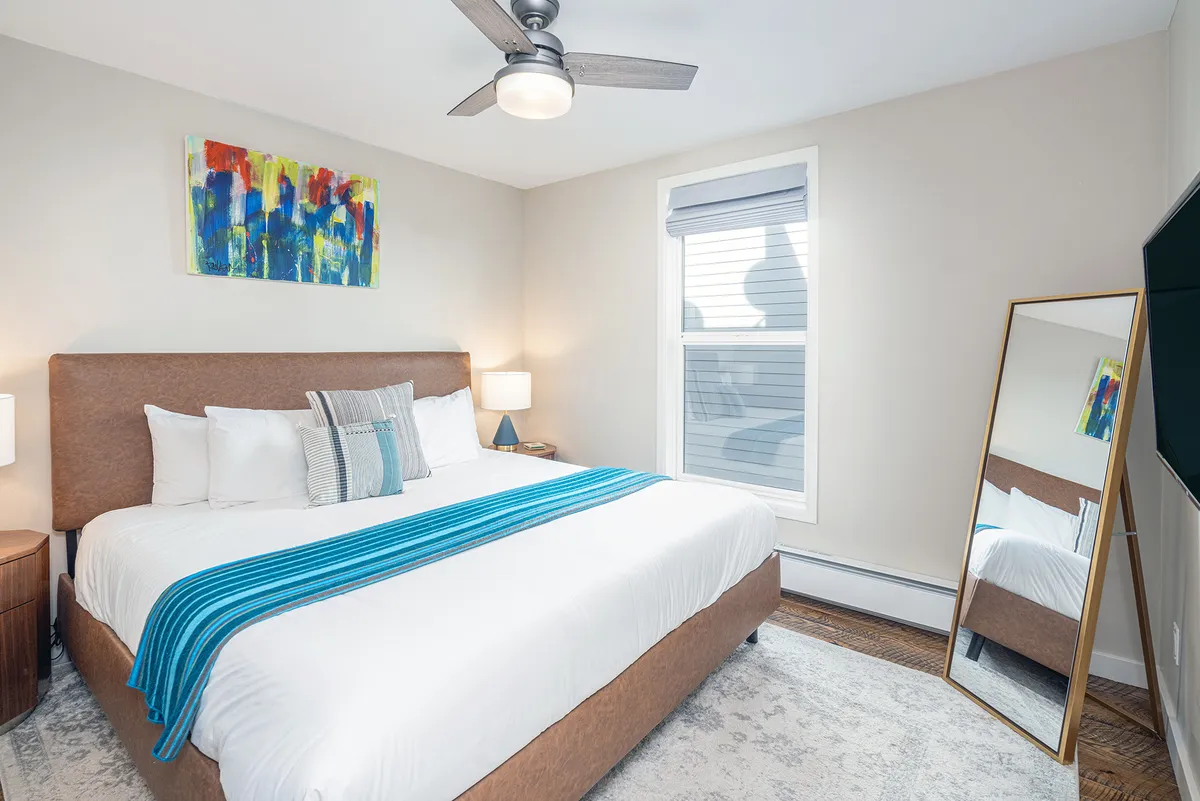 The image showcases a serene bedroom with a large, inviting bed adorned with decorative pillows and a bright blue throw blanket, complemented by modern abstract artwork, a spacious mirror, and a window letting in natural light. The overall feel is tranquil and stylish, highlighting the updated, modern design and cozy atmosphere.