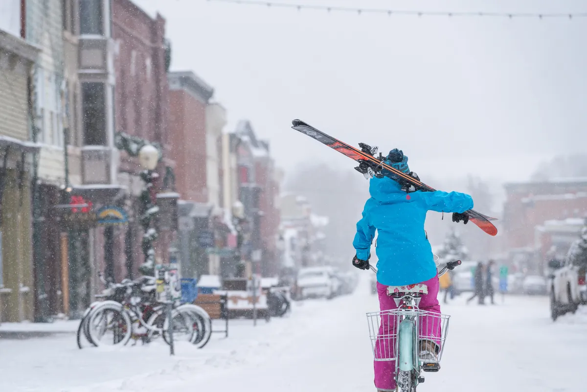 A lively winter street scene in Telluride, featuring a person on a bicycle holding skis over their shoulder, surrounded by snow-covered historic buildings, parked bikes, and people exploring the area. The setting conveys a mix of adventure and local culture, with an inviting, energetic vibe.