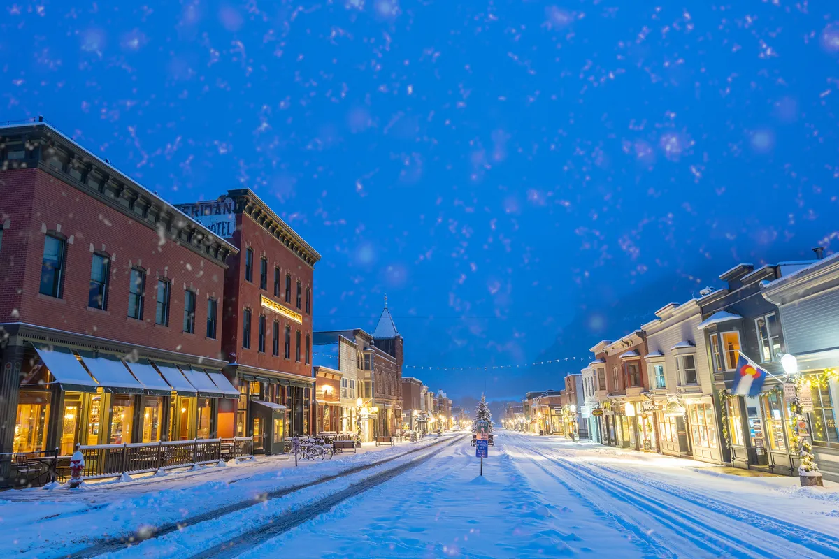 A picturesque winter scene of Telluride's downtown, lined with historic buildings and a snow-covered street, anchored by a large decorated Christmas tree. The street is buzzing with people enjoying holiday shopping, café visits, and the festive atmosphere.