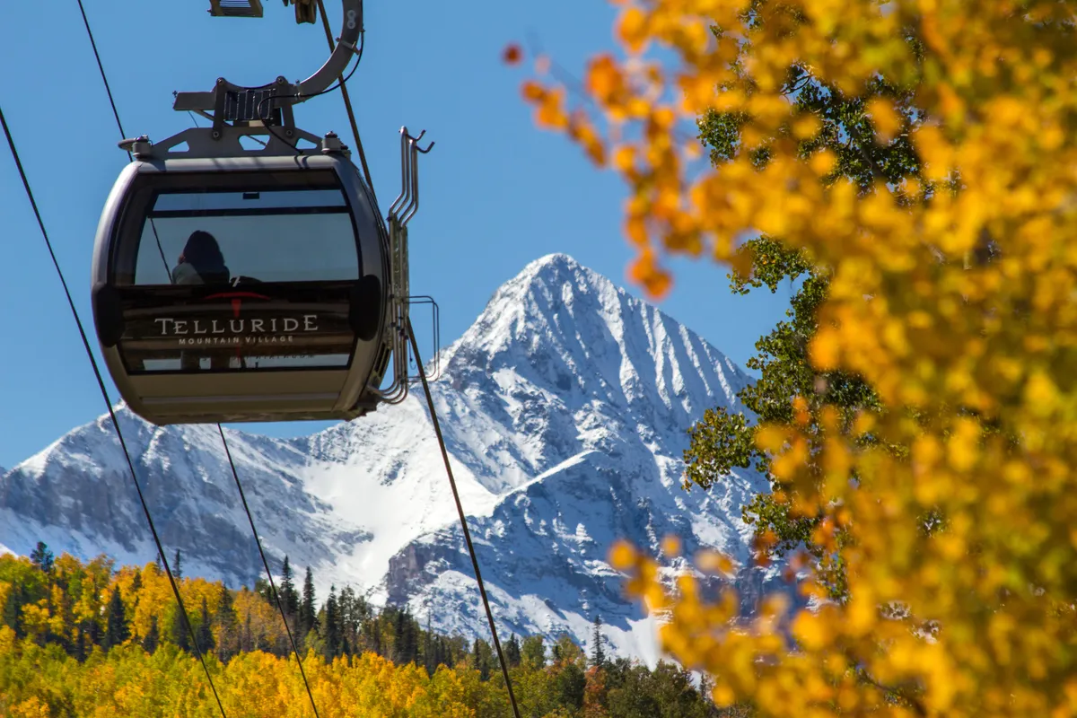 A gondola lift rises up towards majestic, snow-capped mountains while autumn foliage colors the landscape below. The setting hints at both winter and summer activities, with the gondola serving as a gateway to skiing, snowboarding, and hiking trails.