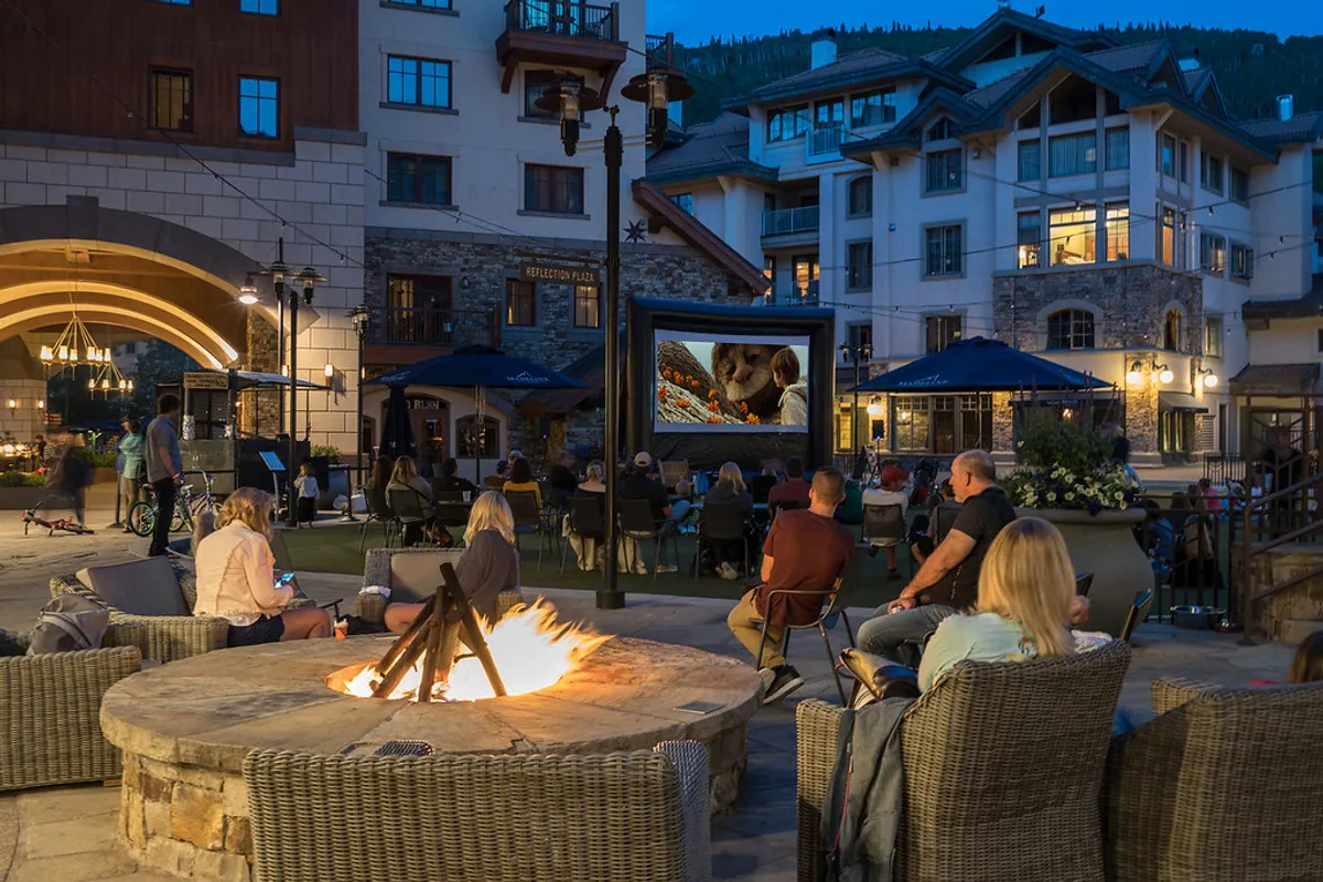 A cozy outdoor gathering around a fire pit with people seated and a movie projected onto an outdoor screen creates a warm, communal vibe. The soft glow of the fire and the sense of togetherness capture a relaxed, inviting atmosphere perfect for unwinding after a day of adventure.